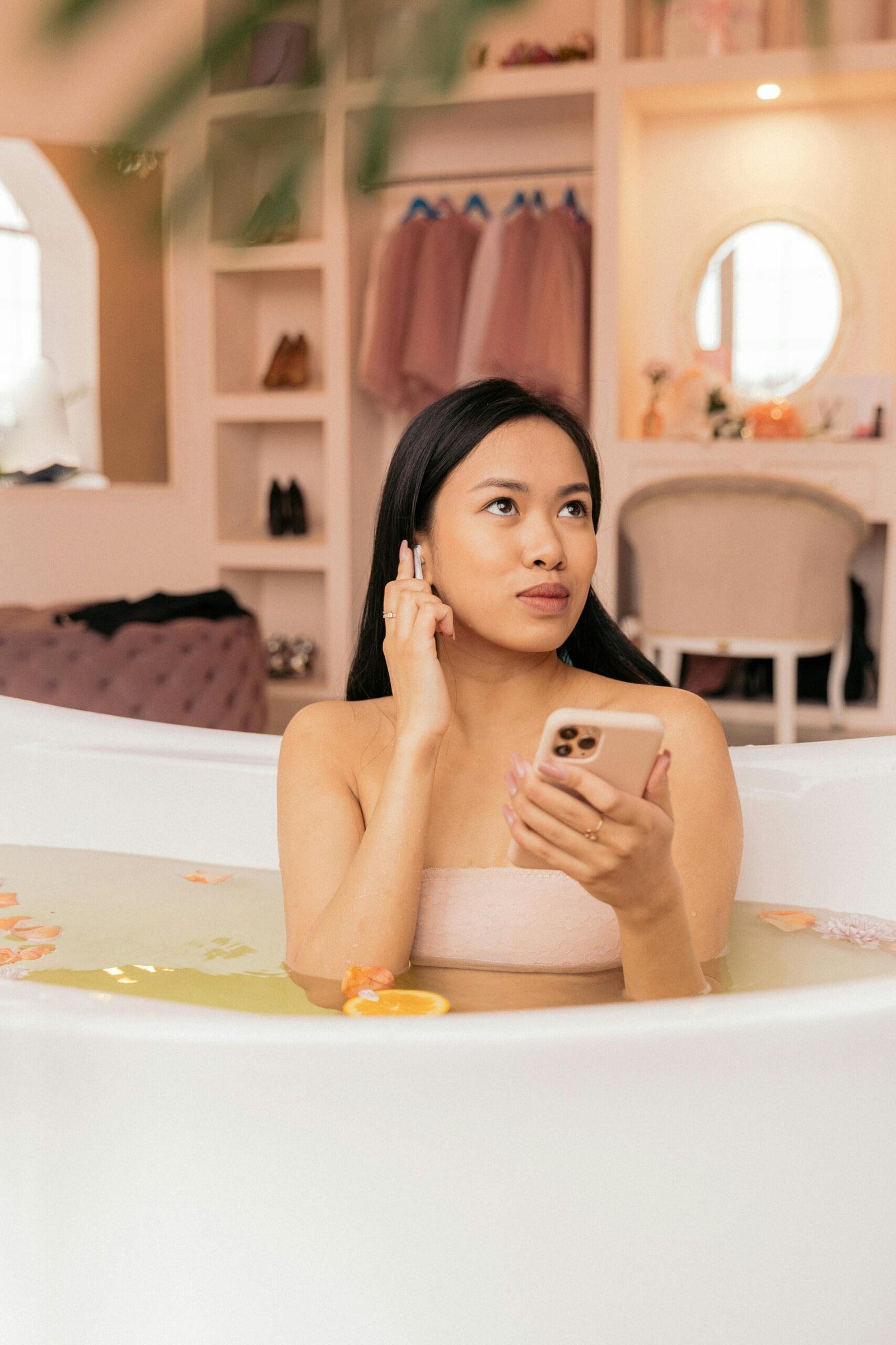 Woman enjoying a calming bath with petals and using a smartphone for relaxation.