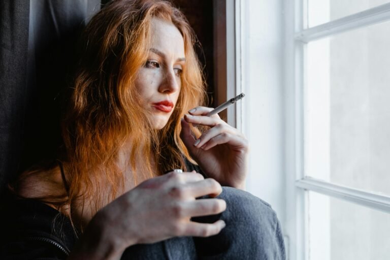 Pensive young woman with red hair holding a cigarette by a window, reflecting indoors.