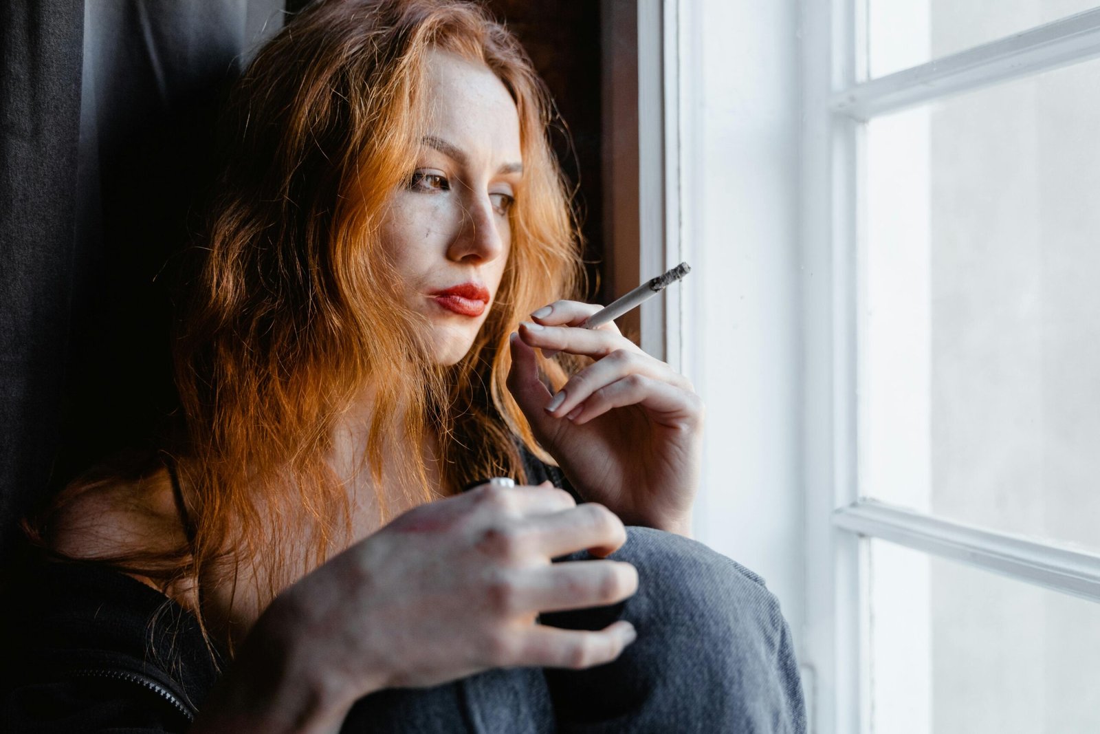 Pensive young woman with red hair holding a cigarette by a window, reflecting indoors.